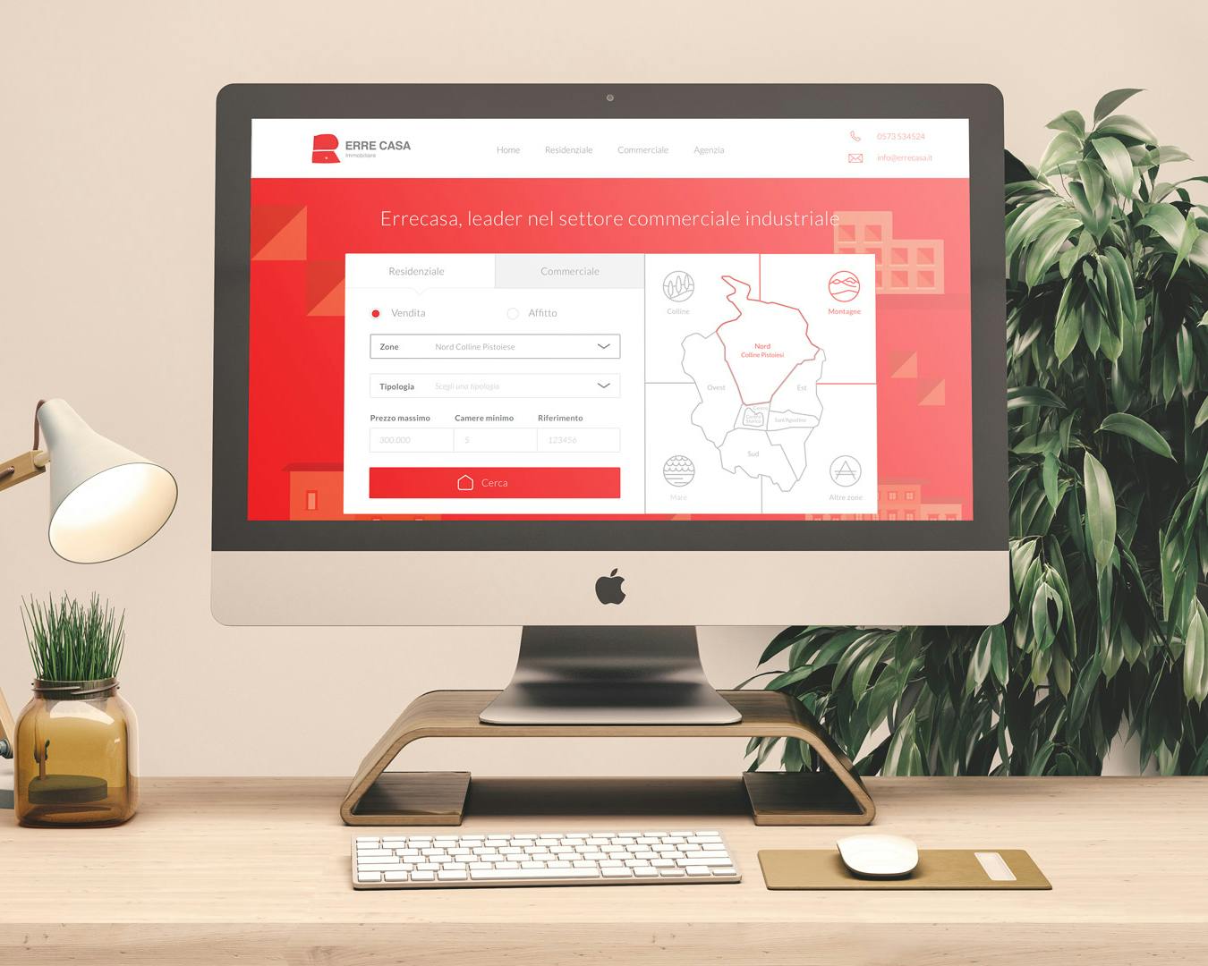 Una scrivania ben organizzata presenta un computer desktop con schermo acceso che visualizza un sito web aziendale dall'interfaccia rossa, accompagnato da una lampada moderna, una pianta e accessori di cancelleria in un ambiente ufficio elegante.