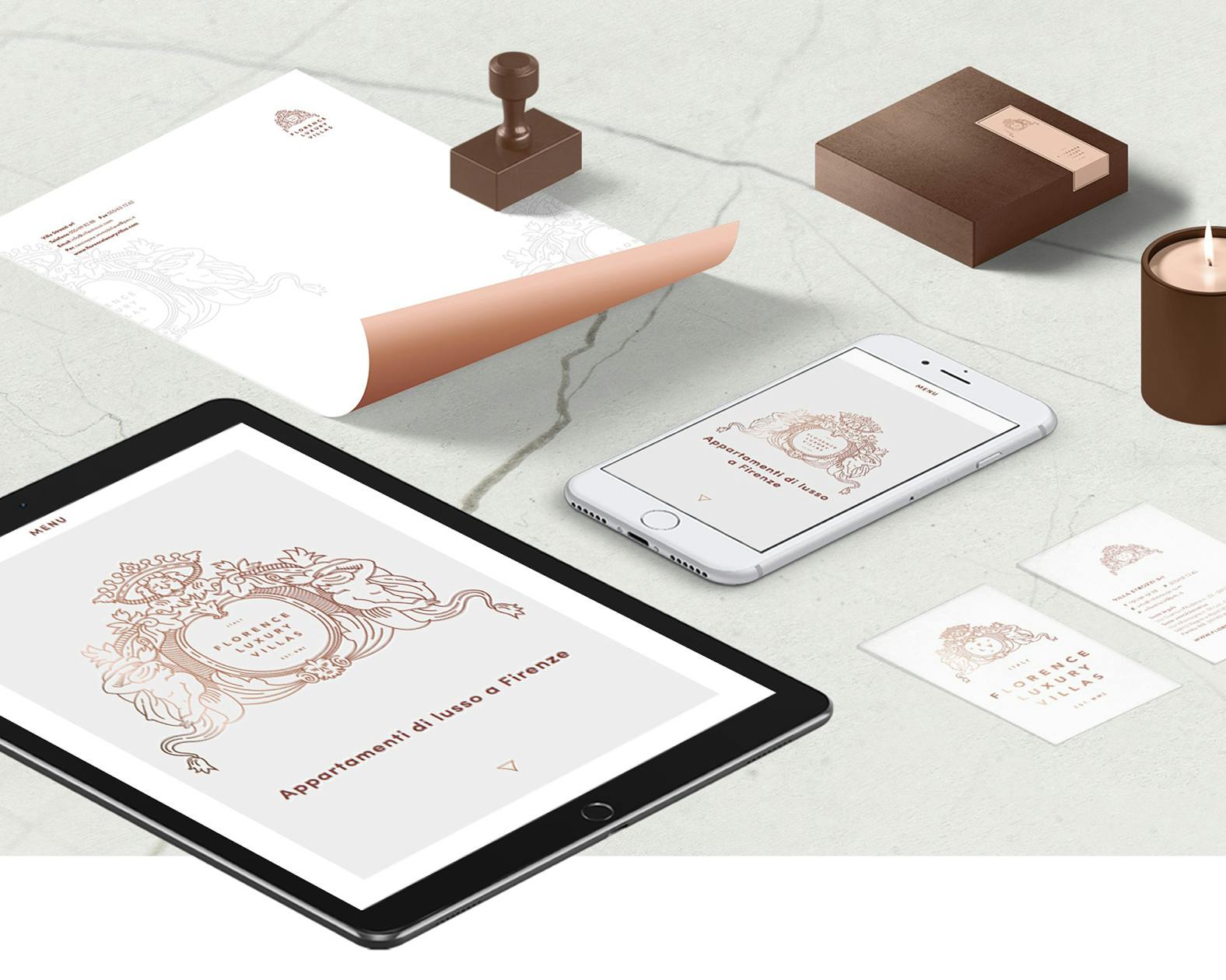 Un set di articoli di cancelleria elegante, tra cui una busta, un timbro, un telefono, un tablet, biglietti da visita e una candela profumata, disposti su una superficie di marmo chiaro.