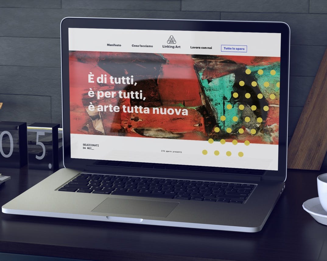 Un laptop è posizionato su una scrivania moderna vicino a un orologio da tavolo e una tazza, con lo schermo che visualizza un sito web colorato e artistico, mentre sullo sfondo si trova una piccola scultura di legno.