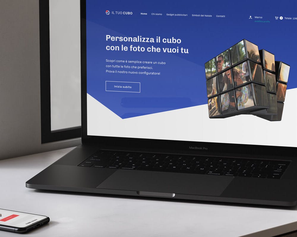Un computer portatile nero con schermo aperto mostra un sito web che offre la personalizzazione di cubi con foto personali, accanto a uno smartphone posizionato su un tavolo di legno chiaro.