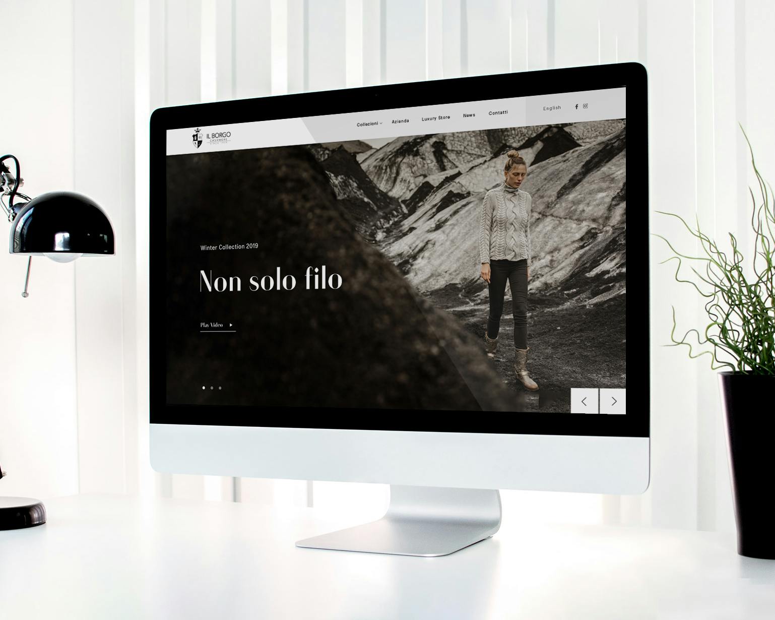 Un moderno computer desktop mostra un sito web di moda con una lampada da scrivania nera e una pianta verde in vaso su una scrivania bianca ben ordinata.