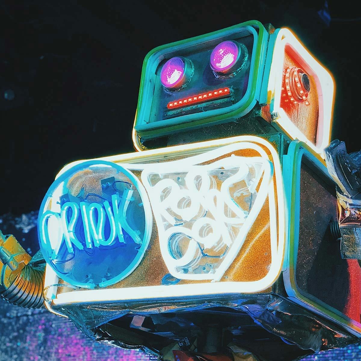 Un robot dallo stile retrò è illuminato da luci al neon colorate, con occhi a forma di obiettivo e dettagli brillanti, posizionato di fronte a uno sfondo scuro e sfocato.