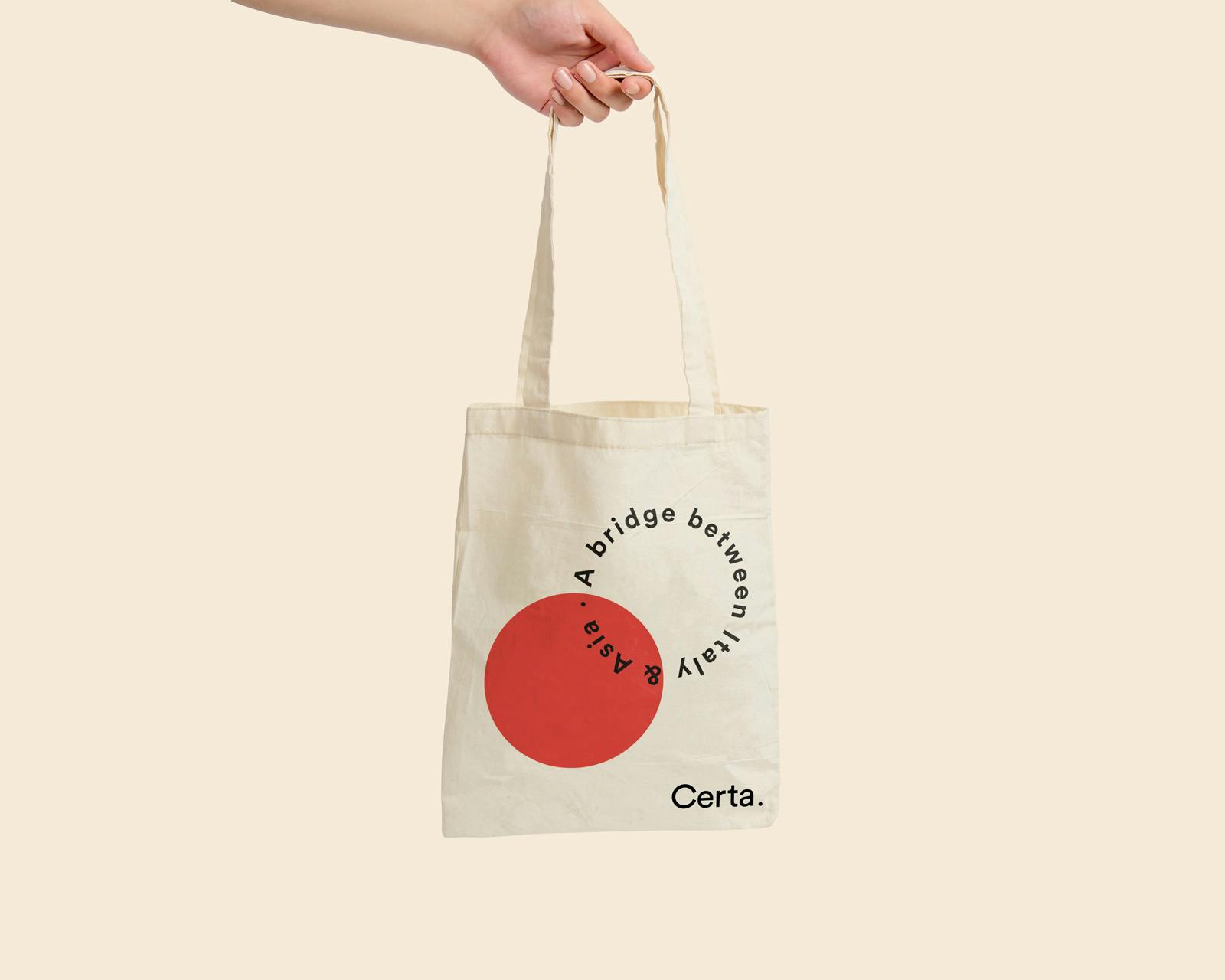 Una mano tiene una borsa di tela beige con un grande cerchio rosso e la scritta "A bridge between Italy & Asia", con il marchio "Certa" nell'angolo in basso a destra.
