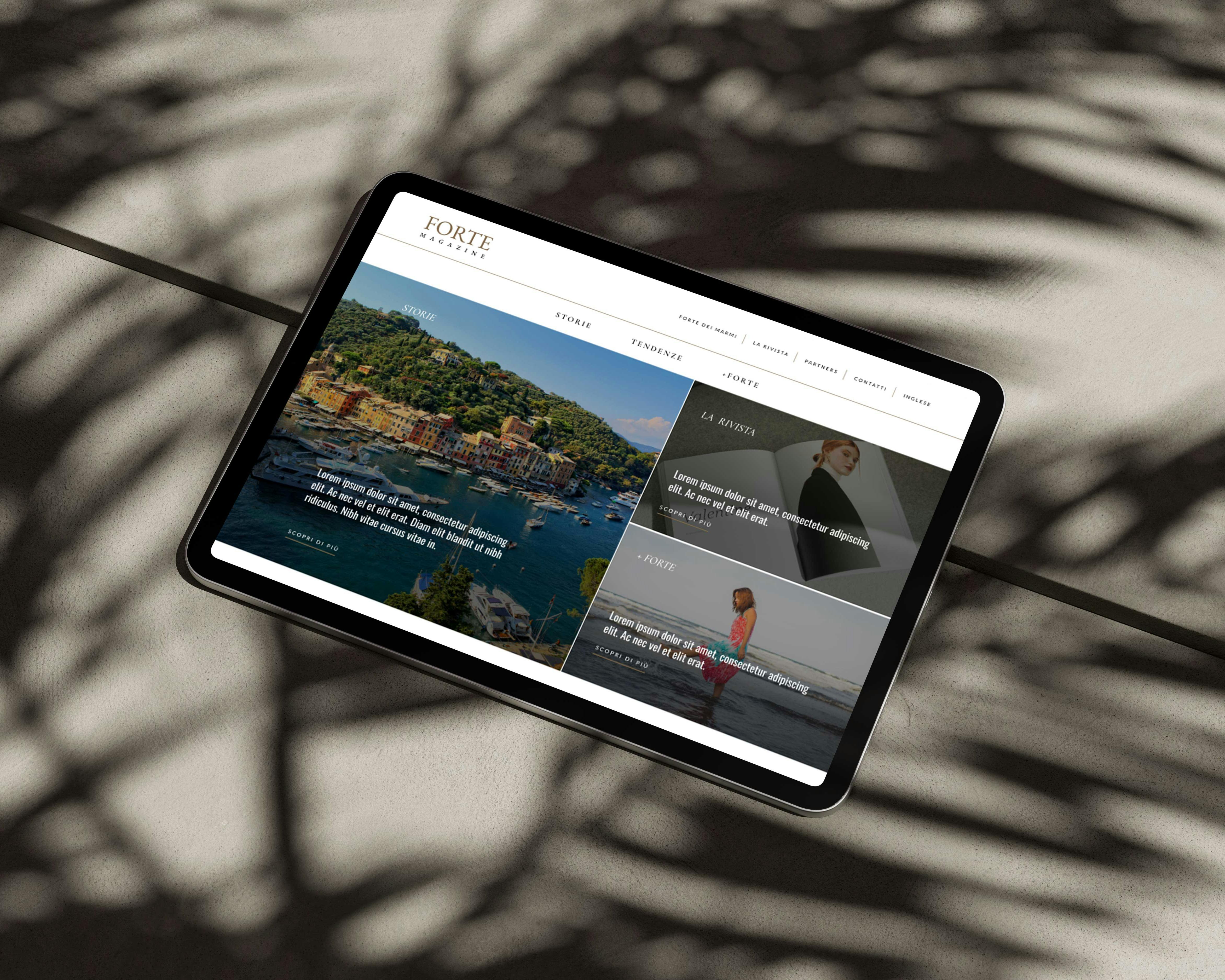 Un tablet nero visualizza un sito Web con immagini di paesaggi costieri colorati, poggiato su una superficie beige con ombre di foglie di palma.
