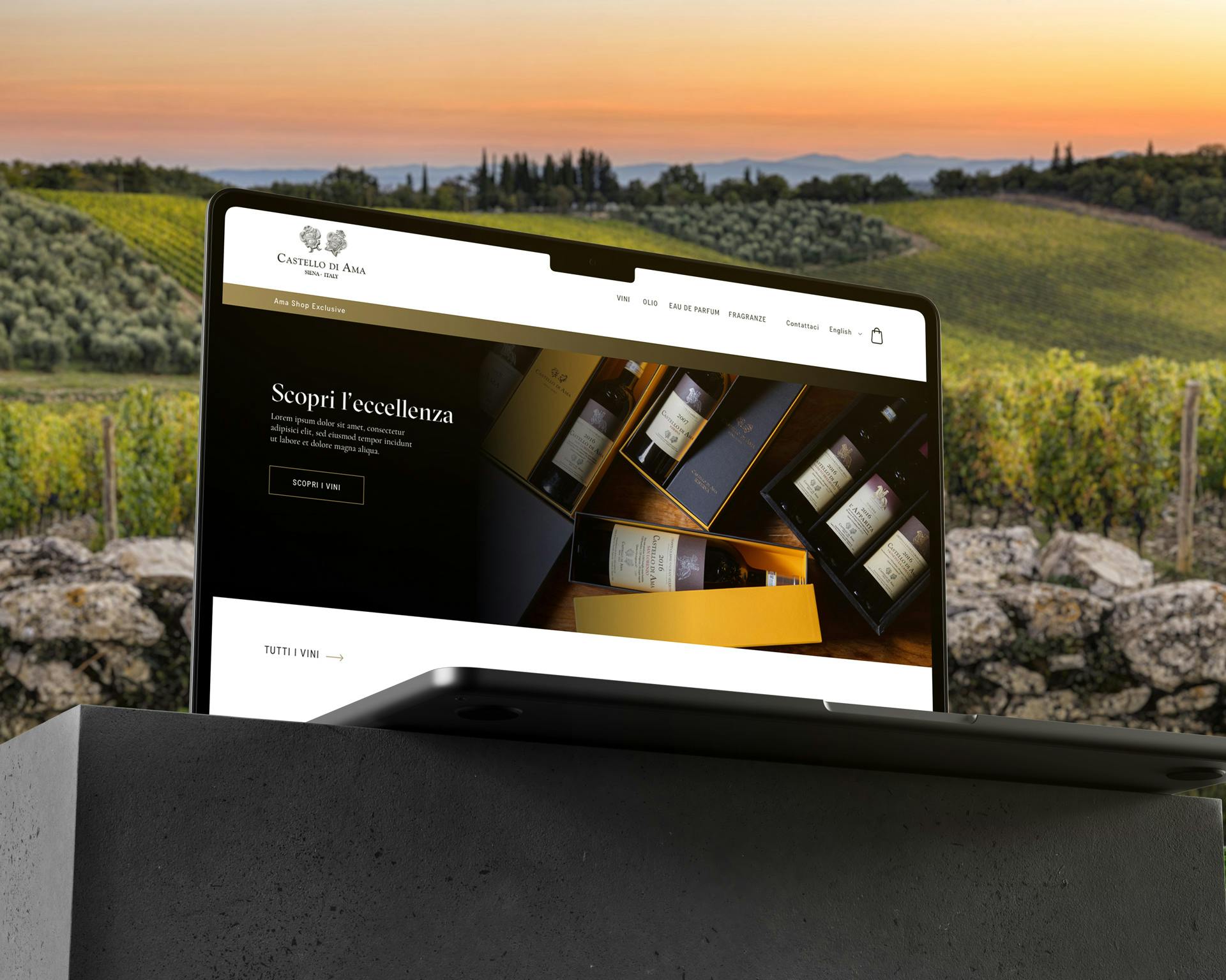 Un laptop aperto mostra un sito web dedicato al vino, posizionato su un tavolo di pietra con una pittoresca vigna verde e un tramonto all'orizzonte.