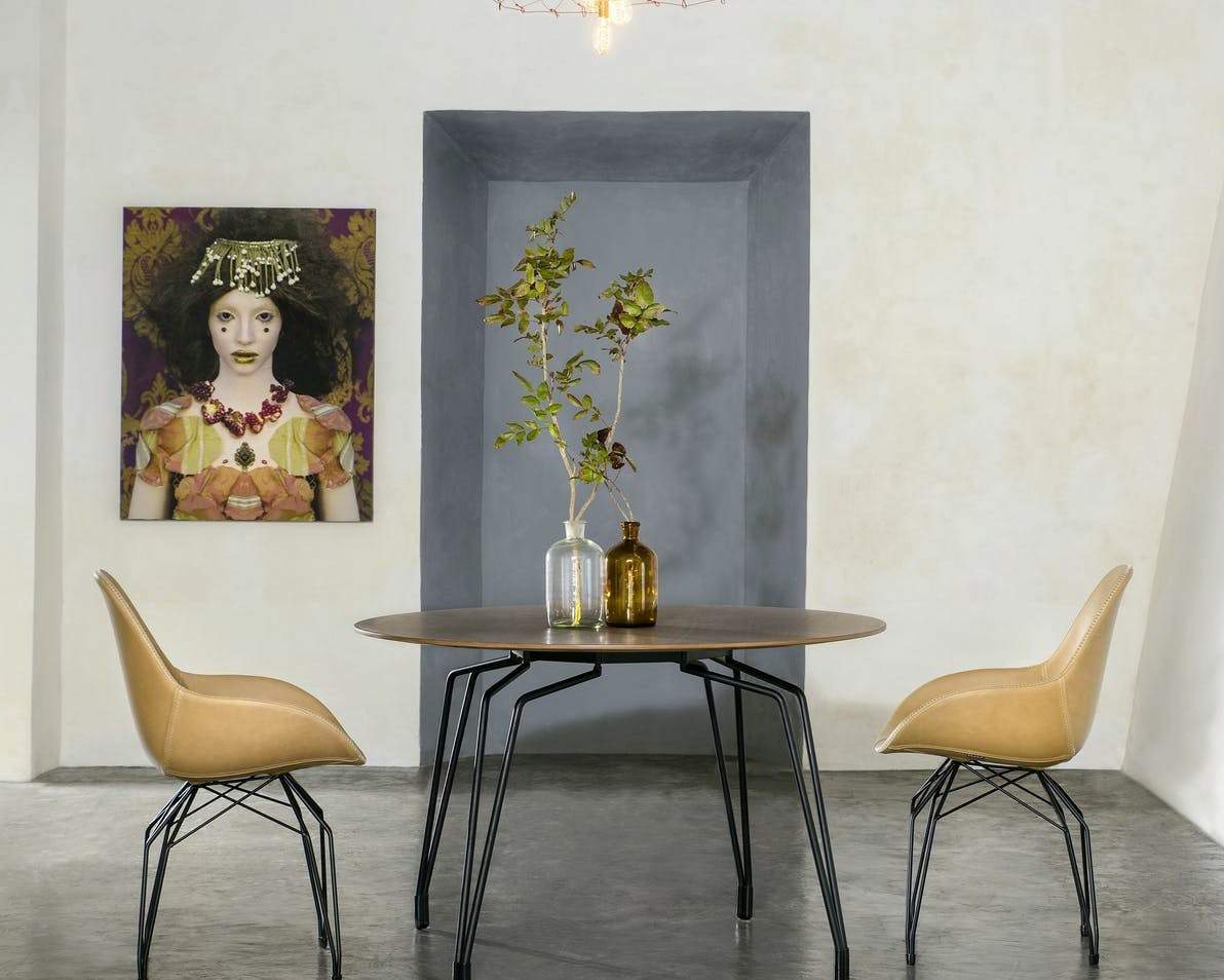 Due sedie moderne color beige con gambe in metallo nero circondano un tavolo rotondo in legno, sopra il quale si trova un vaso con rami verdi; sullo sfondo, un grande quadro decorativo di una donna adorna la parete accanto a una lampada a sospensione.