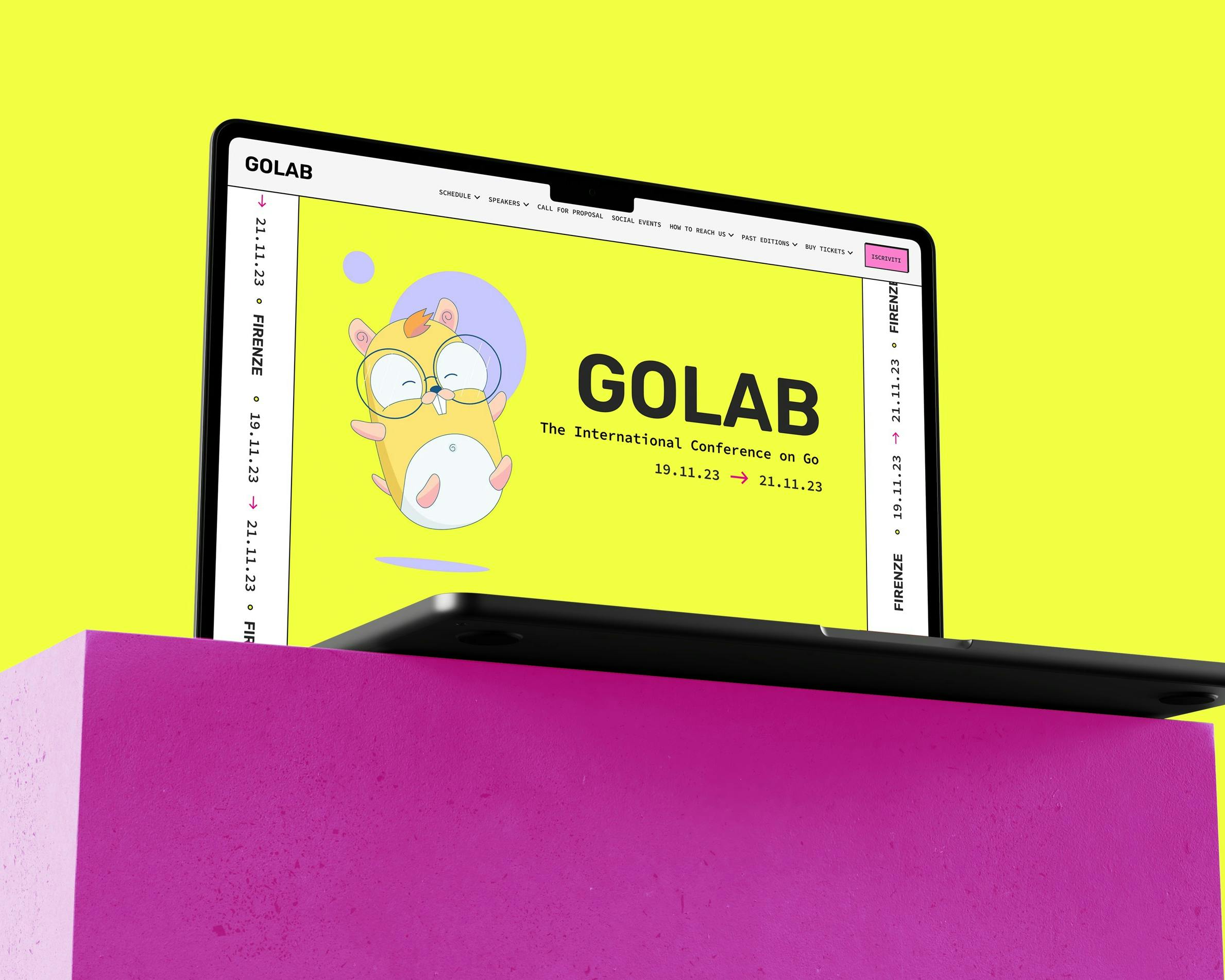 Un laptop aperto mostra una schermata gialla con il logo di GO e un testo che annuncia una conferenza internazionale sul linguaggio Go, sopra un cubo rosa acceso.