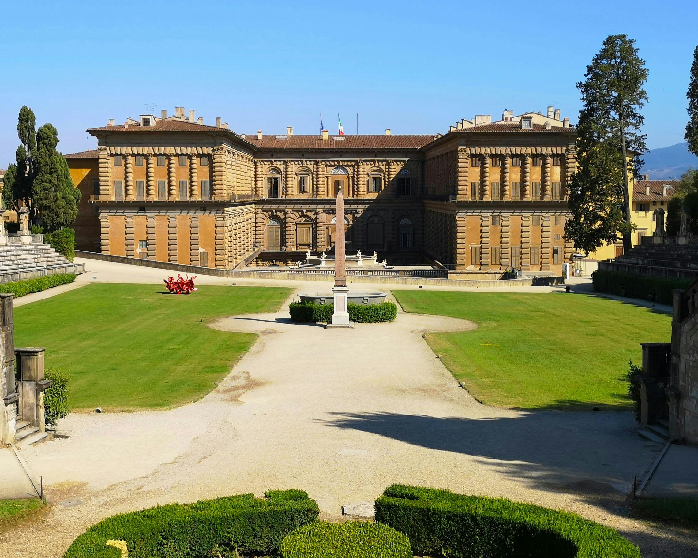 Il Palazzo Pitti a Firenze è visto dal giardino, con un viale che conduce all'imponente facciata storica, circondata da prati ben curati e alberi rigogliosi.