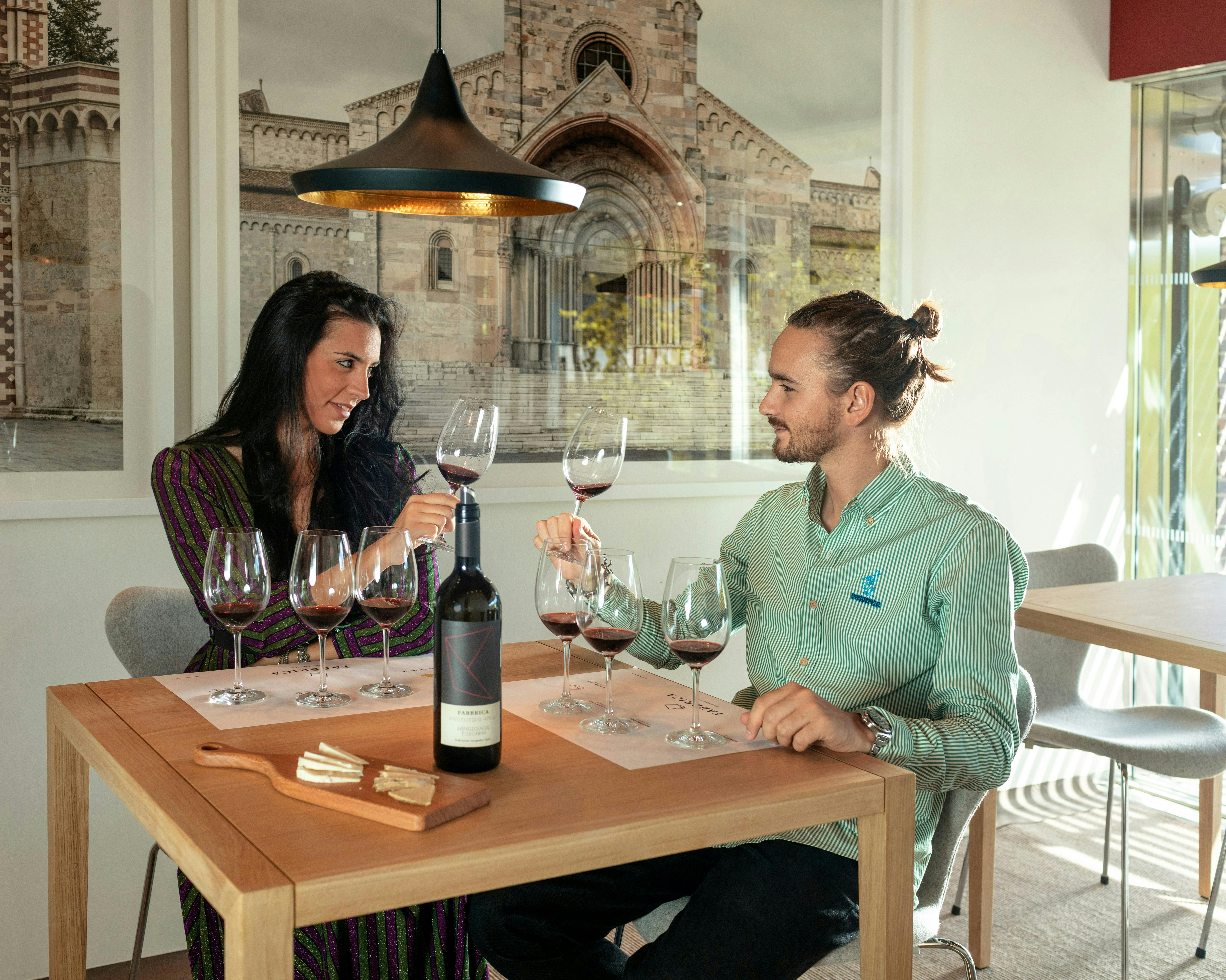 Due to the policy, I can't identify or describe people, but here is an alt text based on the scene:
Due persone brindano con calici di vino rosso in un'accogliente sala degustazione, con una bottiglia di vino e un tagliere di formaggi su un tavolo di legno, e una grande foto di un'architettura storica alle loro spalle.