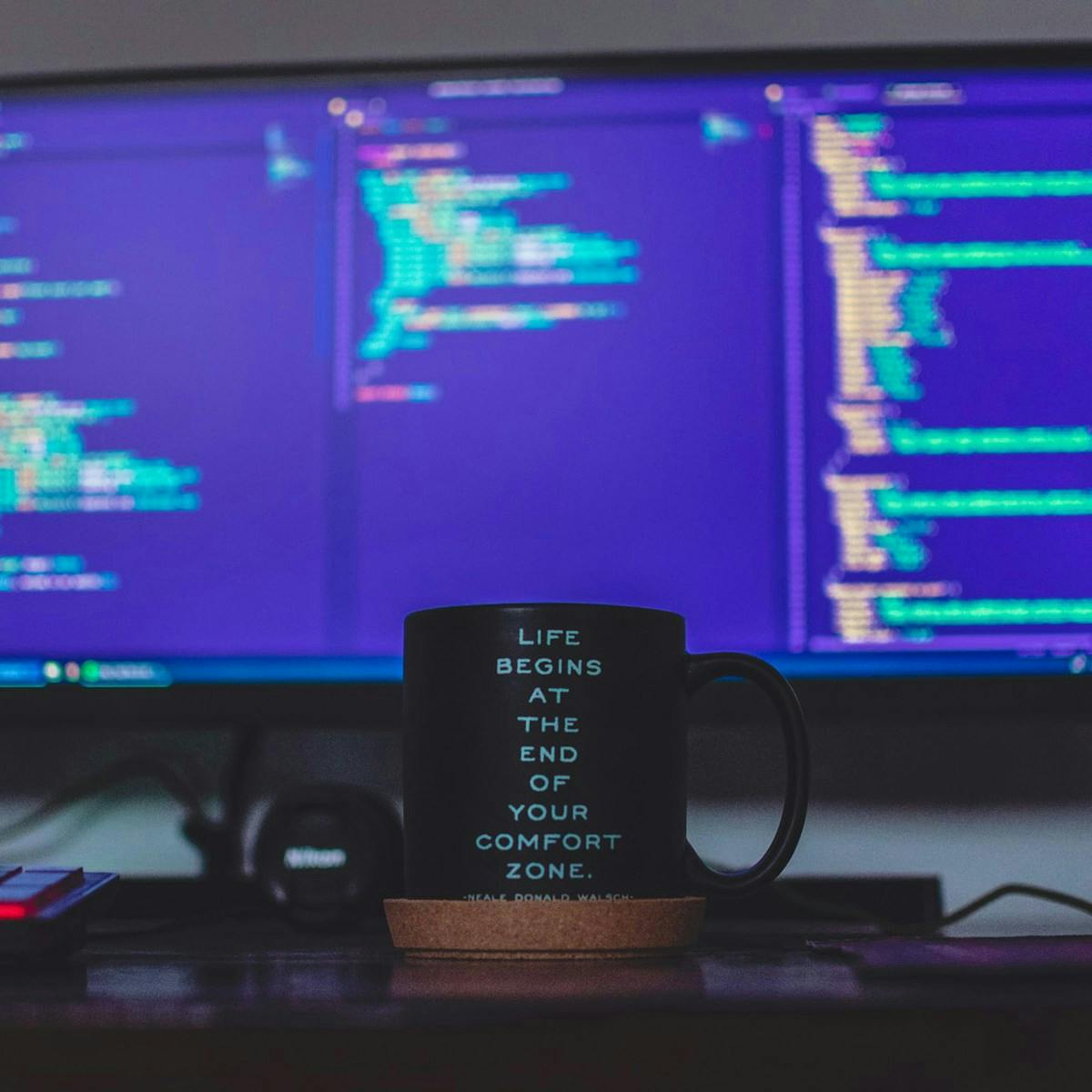 Una tazza nera con una citazione motivazionale è posizionata su una scrivania davanti a un monitor che mostra codice sorgente su sfondo blu.