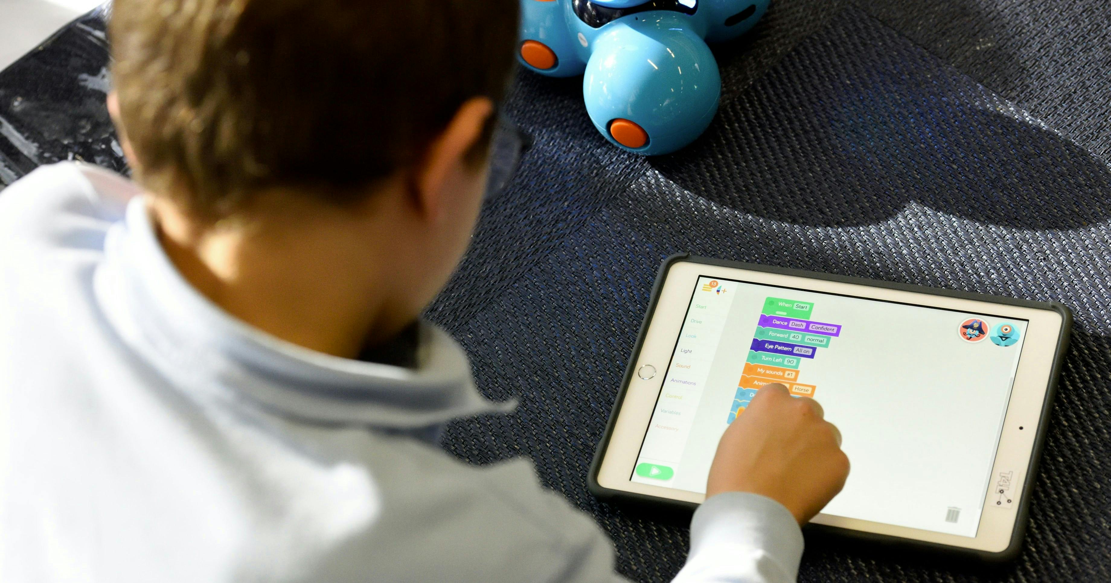 Un bambino concentra la sua attenzione su un tablet mentre interagisce con un'app di programmazione, con un robot giocattolo blu posizionato sullo sfondo, illustrando l'apprendimento tecnologico e l'educazione STEM per bambini.