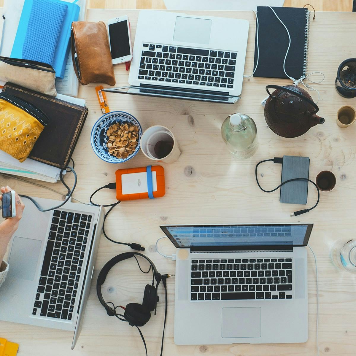 Una scrivania vista dall'alto con tre laptop aperti, circondati da quaderni, un telefono, cuffie, un disco rigido esterno arancione, una tazza di tè, una teiera e varie borse, creando un'atmosfera di lavoro collaborativo o di studio.