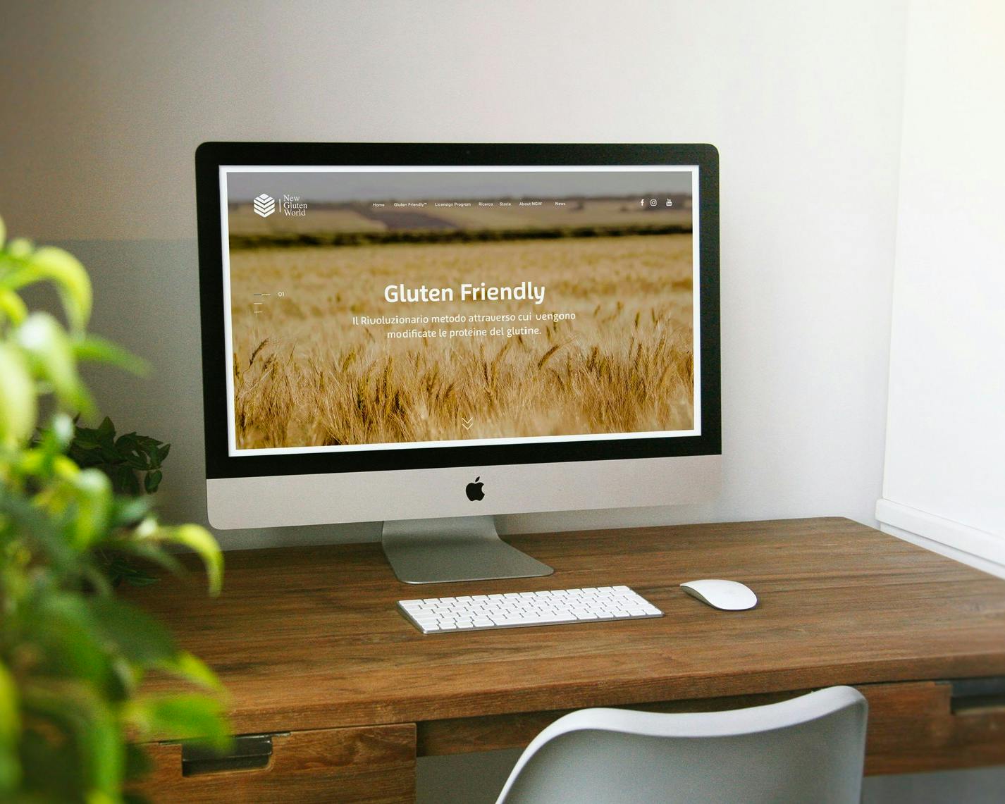 Una scrivania in legno con un computer iMac che mostra una pagina web intitolata "Gluten Friendly", circondata da una pianta verde sul lato sinistro e una sedia bianca di fronte.