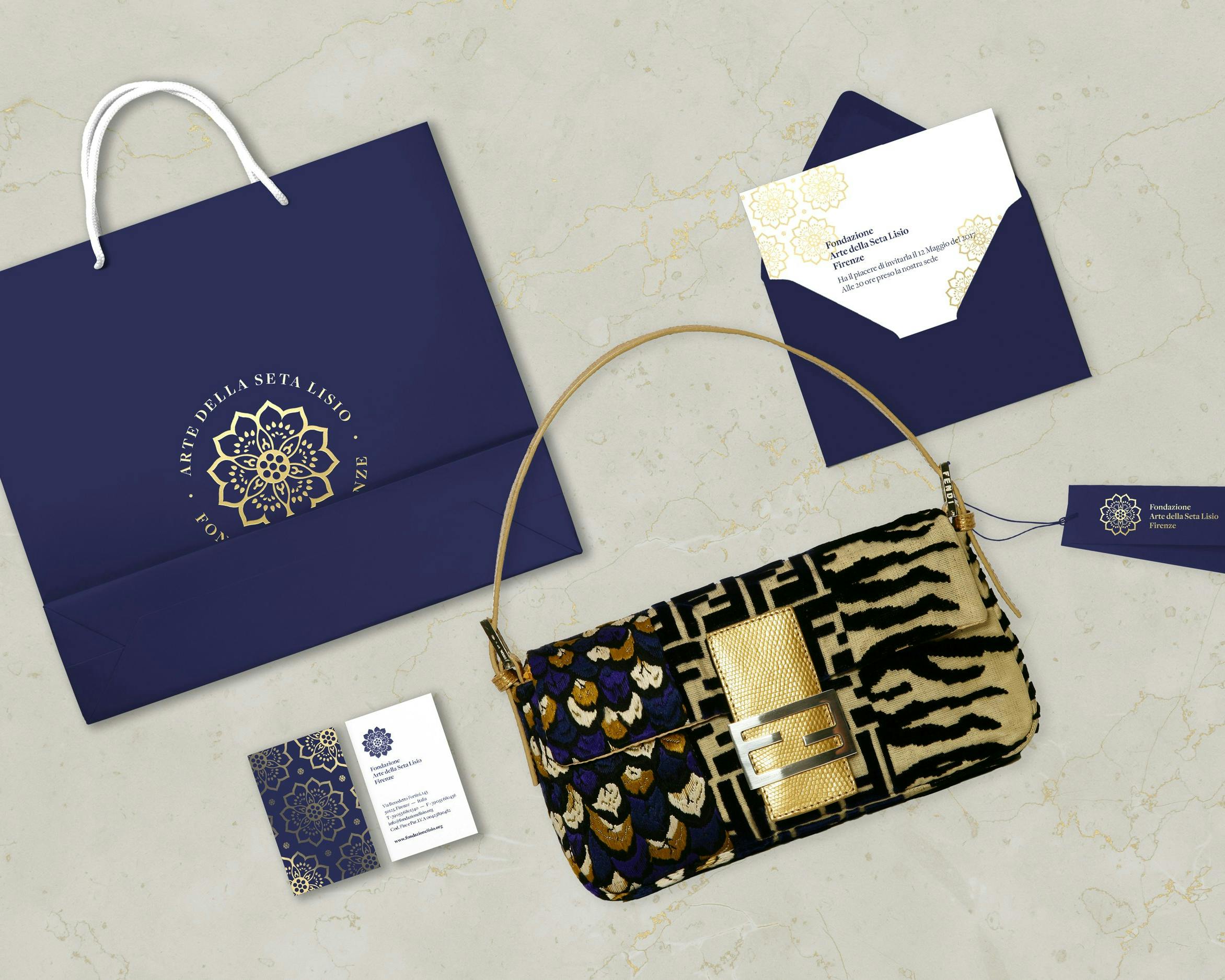 Una borsa a tracolla di design animalier con dettagli dorati è poggiata su una superficie marmorea accanto a una busta regalo blu e bianca, biglietti da visita coordinati e una busta decorata con motivi dorati.