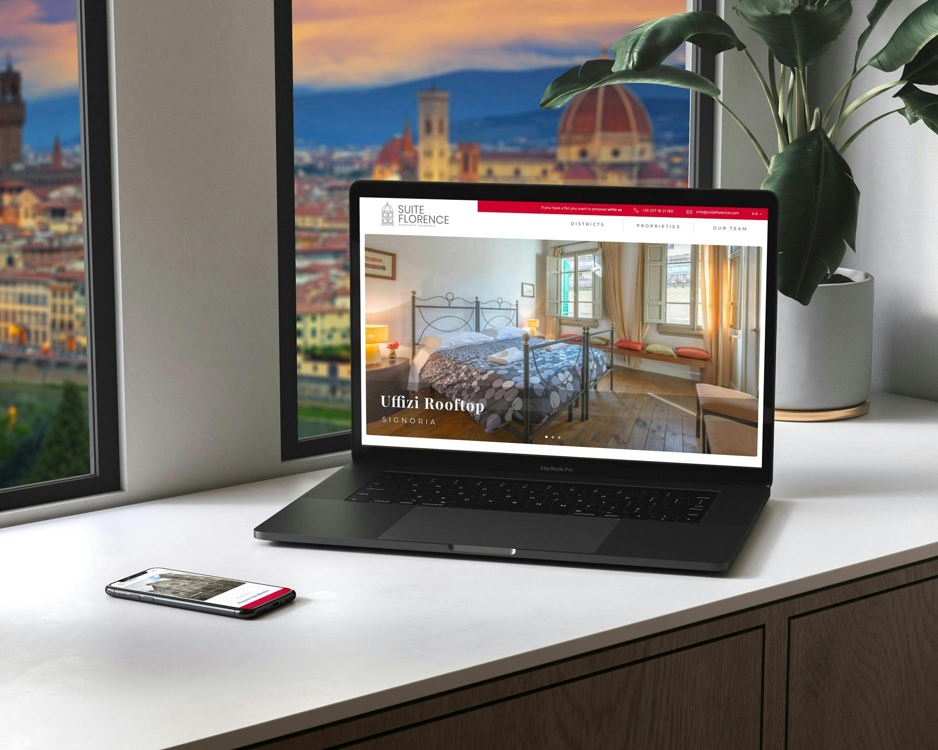 Una scrivania moderna con un laptop che visualizza un sito web di un hotel a Firenze, Italia, affiancato da uno smartphone, davanti a una finestra che offre una vista panoramica della città con il Duomo in lontananza.