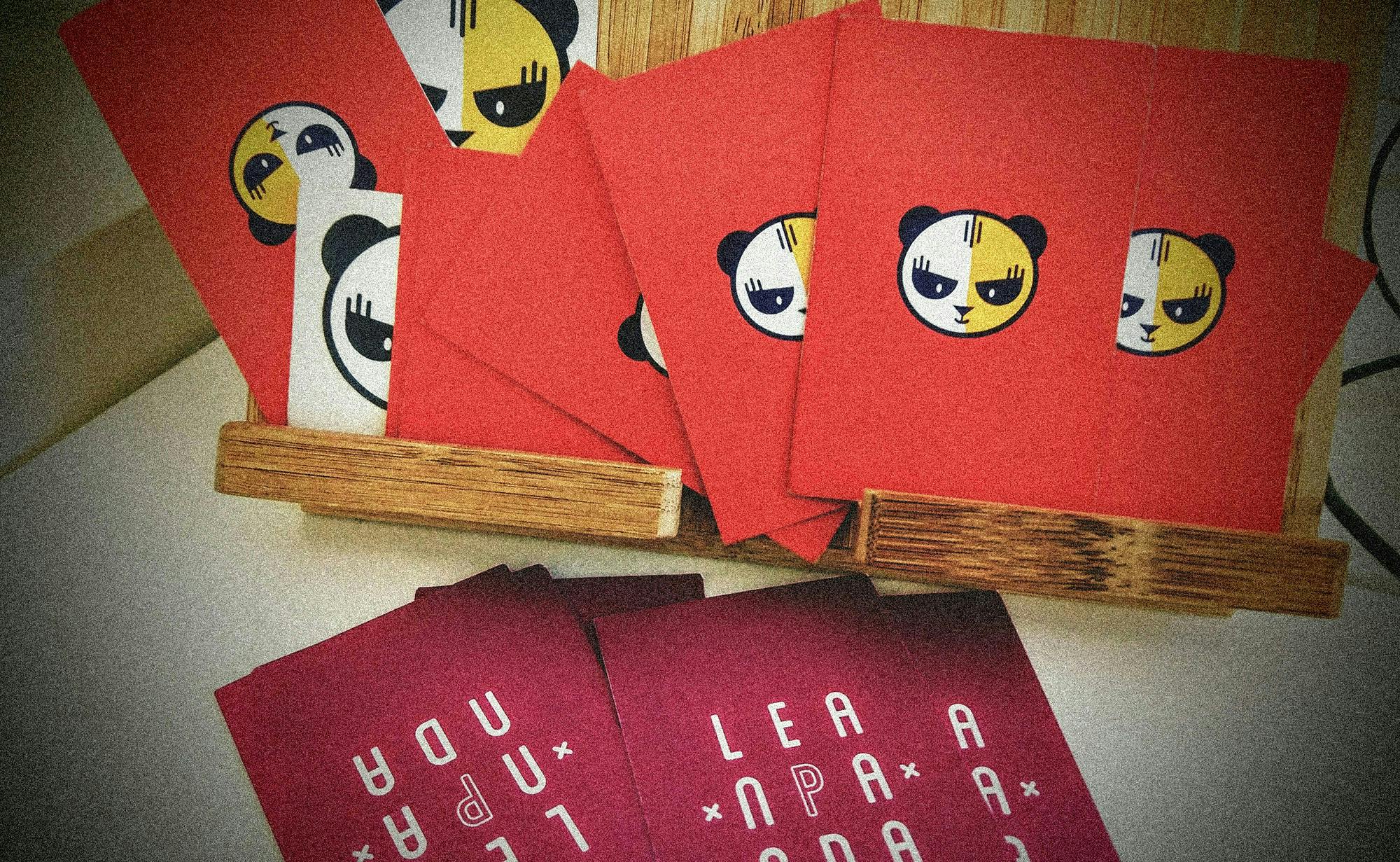 Un set di carte rosse con illustrazioni di panda stilizzati, posizionate su un supporto di legno e accompagnate da carte aggiuntive con scritte artistiche.