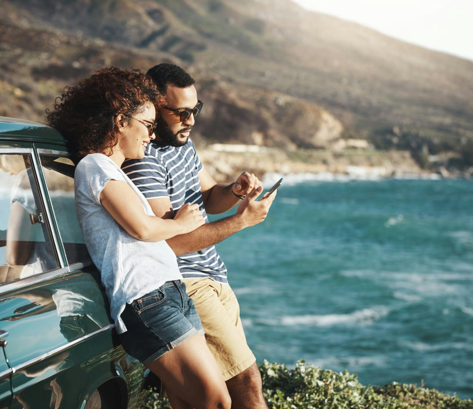 Couple looking at their phone while leaning against their car parked near the ocean