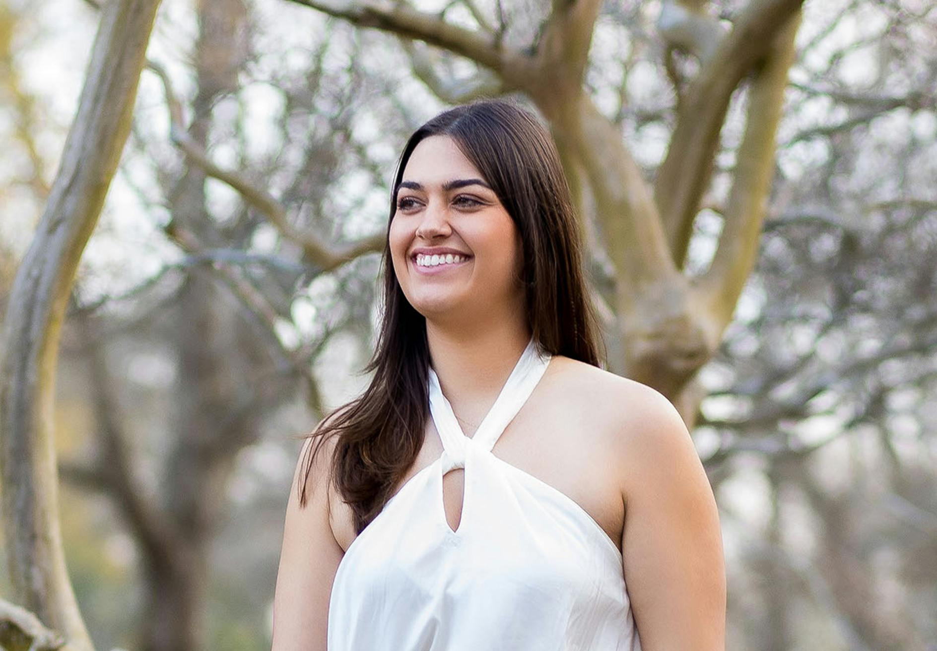 smiling woman in white top standing in front of trees