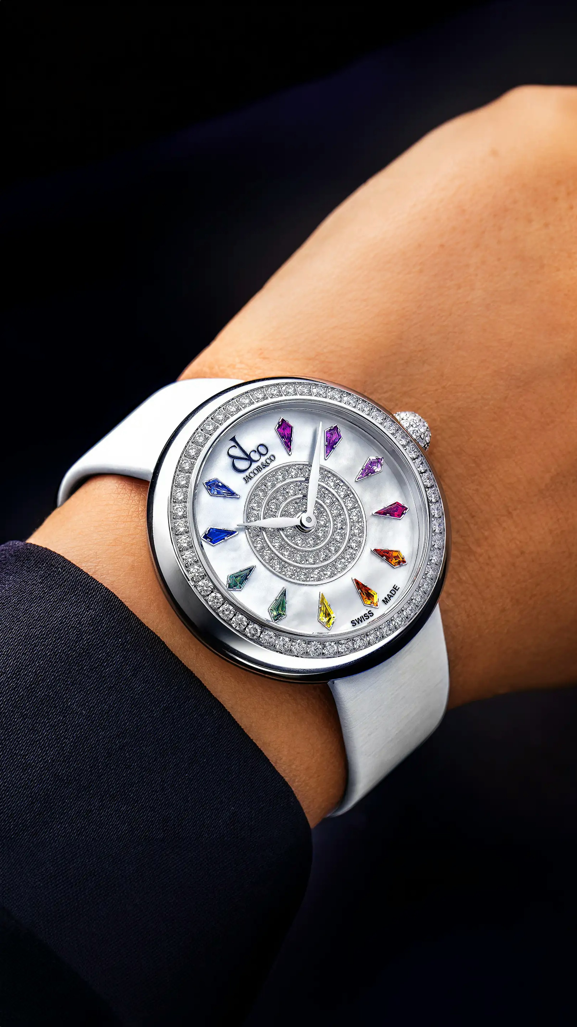 The stainless steel, rainbow Brilliant Watch, with multicolored sapphire hour markers. Worn on the left wrist.