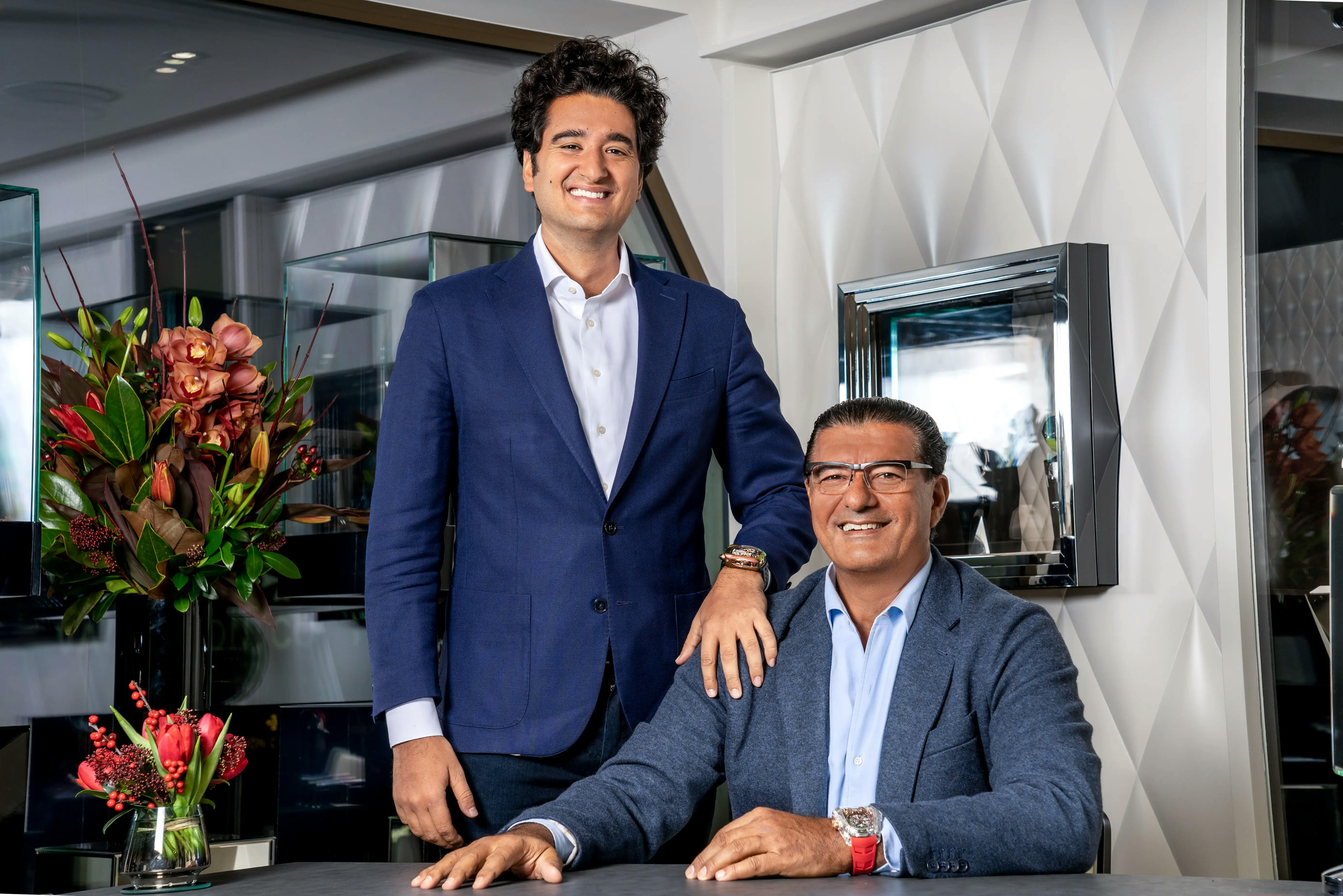 An image of two men in suits. One standing and one sitting behind a table.
