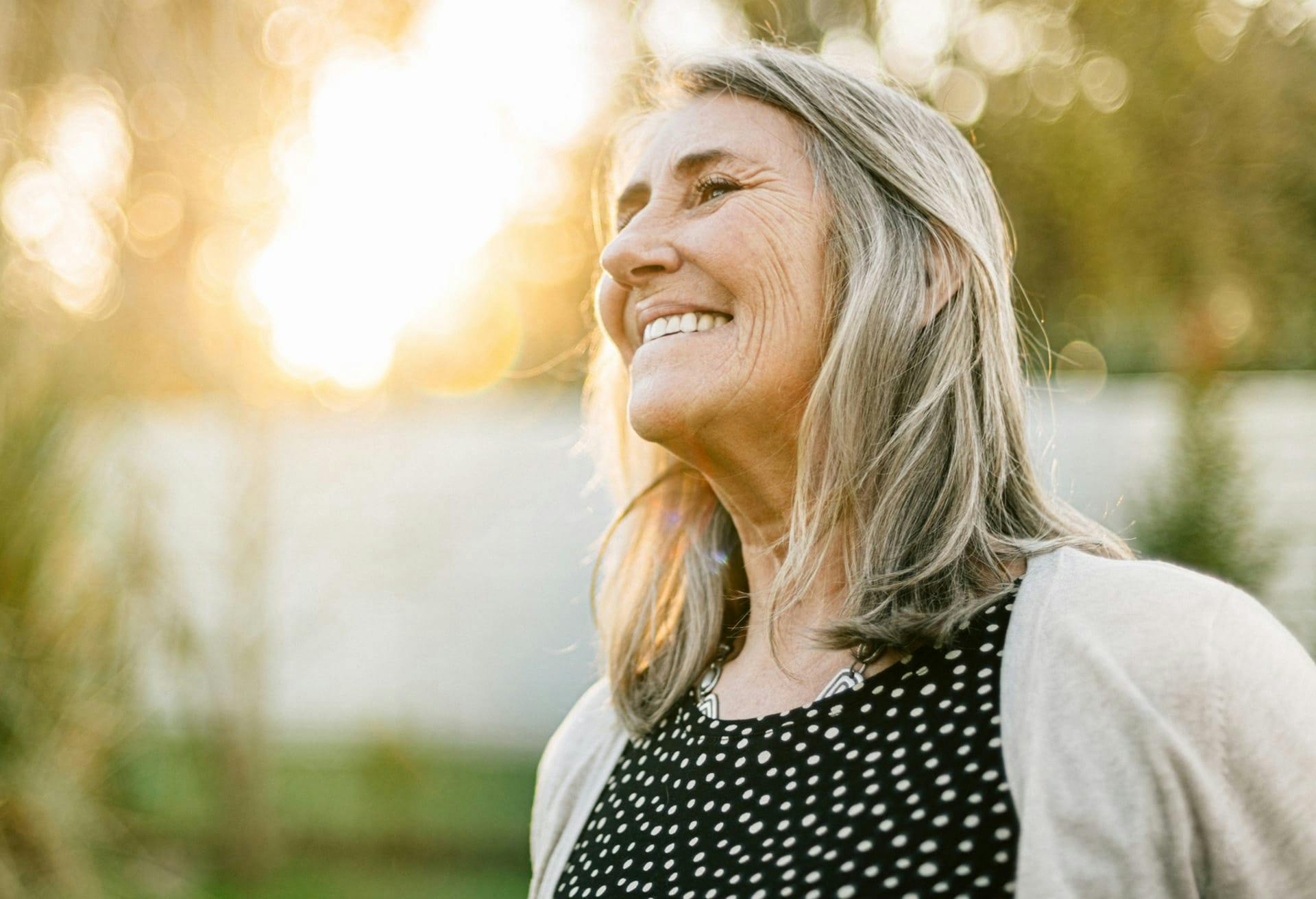 smiling woman with gray hair and black polka dot dress in front of a white fence