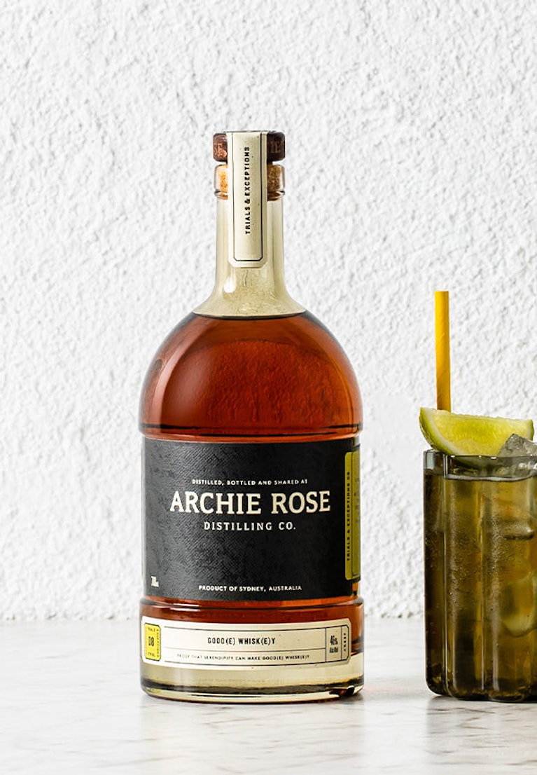 archie-rose-goode-whiskey-front-on-full-bottle-shot-on-table-beside-whisky-cocktail-and-fresh-limes
