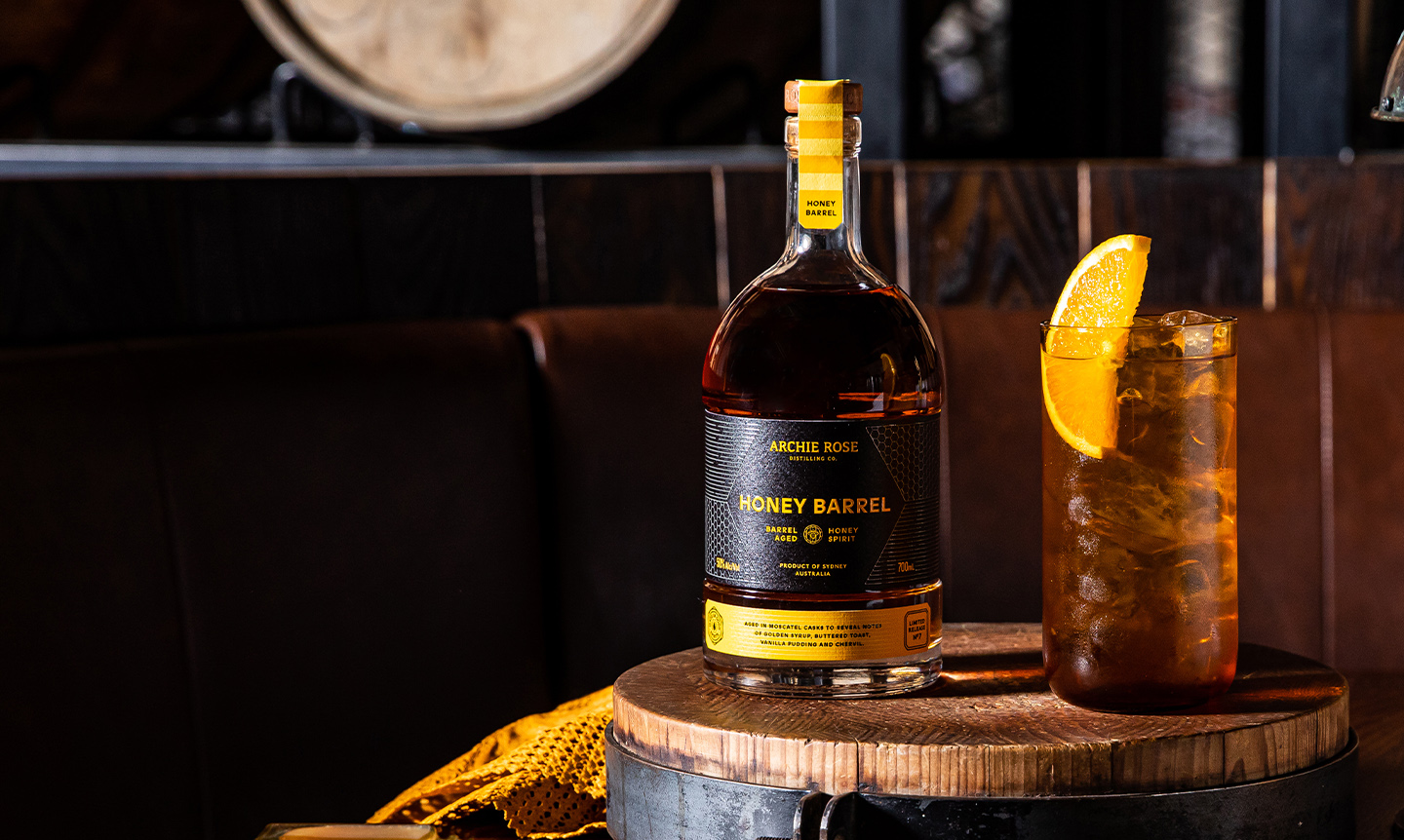 archie-rose-honey-barrel-aged-spirit-front-on-bottle-shot-with-cocktail-in-tall-glass-with-lemon-wedge