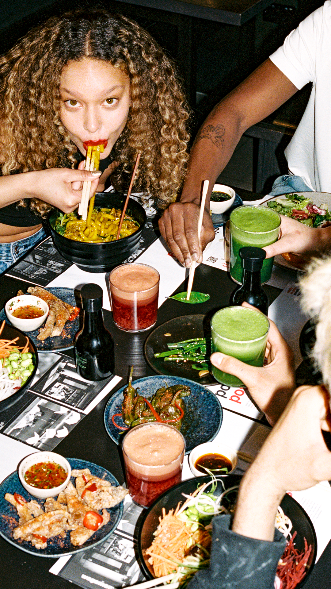 a woman eating noodles with chopsticks sat at a table of food and drink