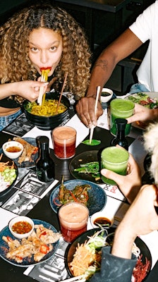 a woman eating noodles with chopsticks sat at a table of food and drink