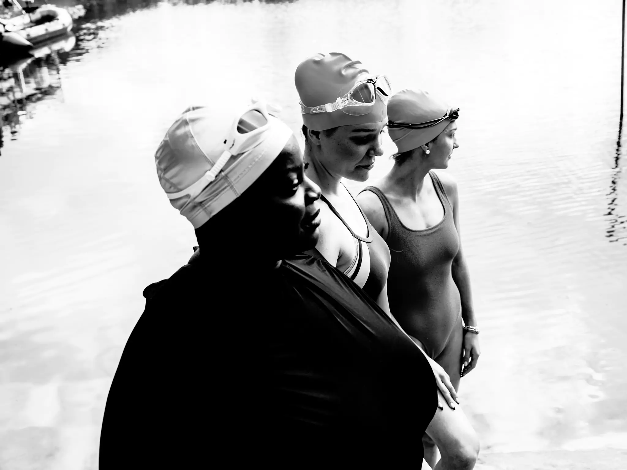 three people stood by water in swimming suits, swimming hats and goggles