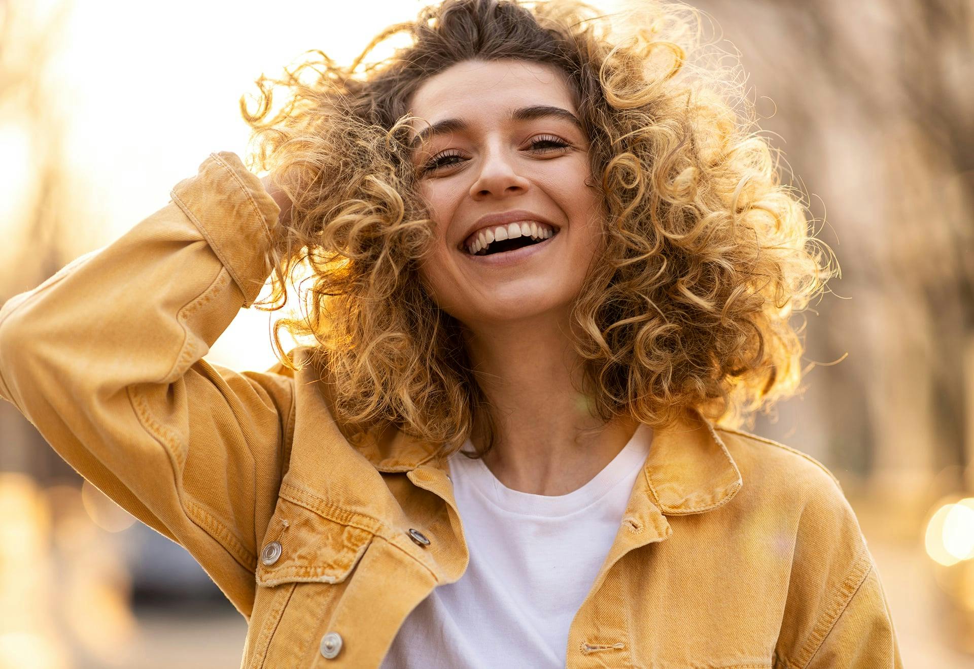 Woman with curly blonde hair smiling, with the sun behind her