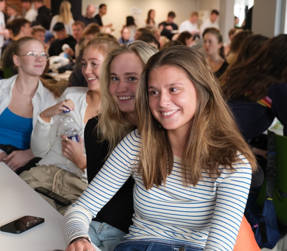 tre piger sidder smilende i kantinen på RKS blandt en masse andre elever