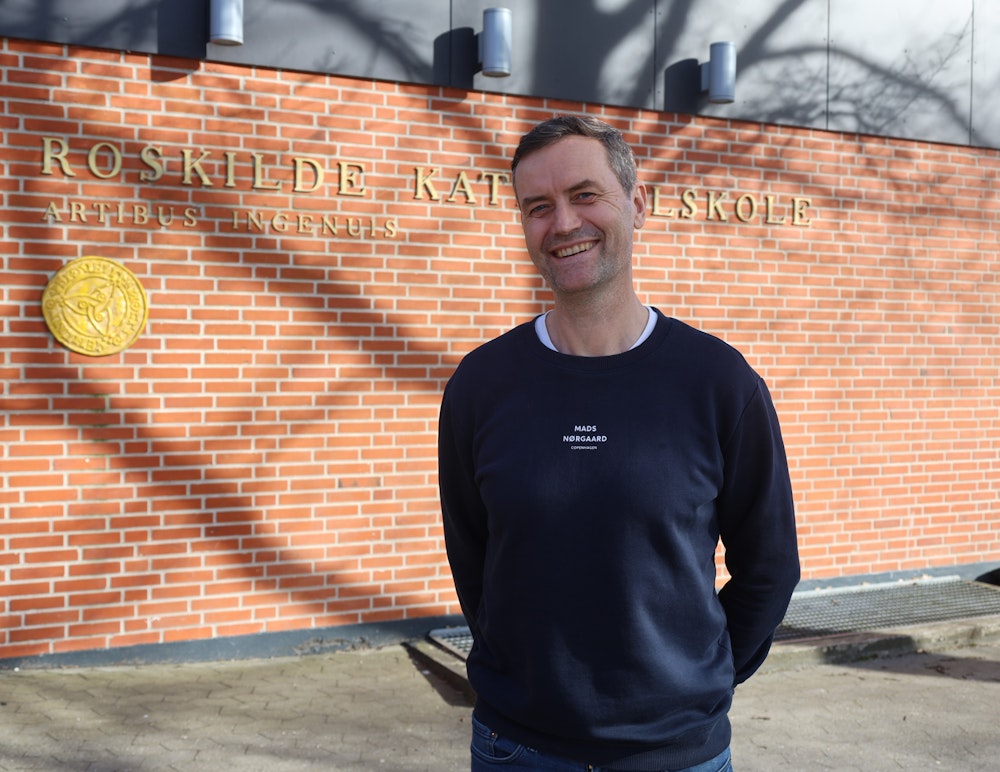 Rektor Thomas Stilling står smilende foran gymnasiets logo ved hovedindgangen