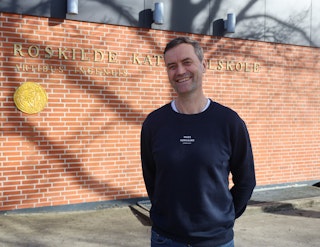 Rektor Thomas Stilling står smilende foran gymnasiets logo ved hovedindgangen