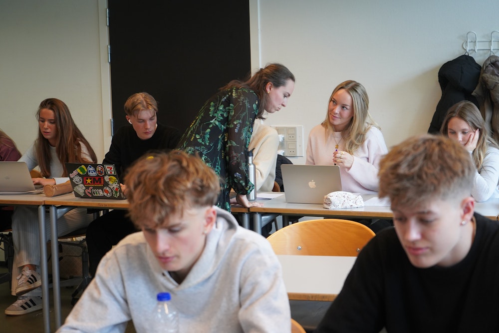 Et klasseværelse, hvor en lærer forklarer noget til en elev, der sidder med åben pc