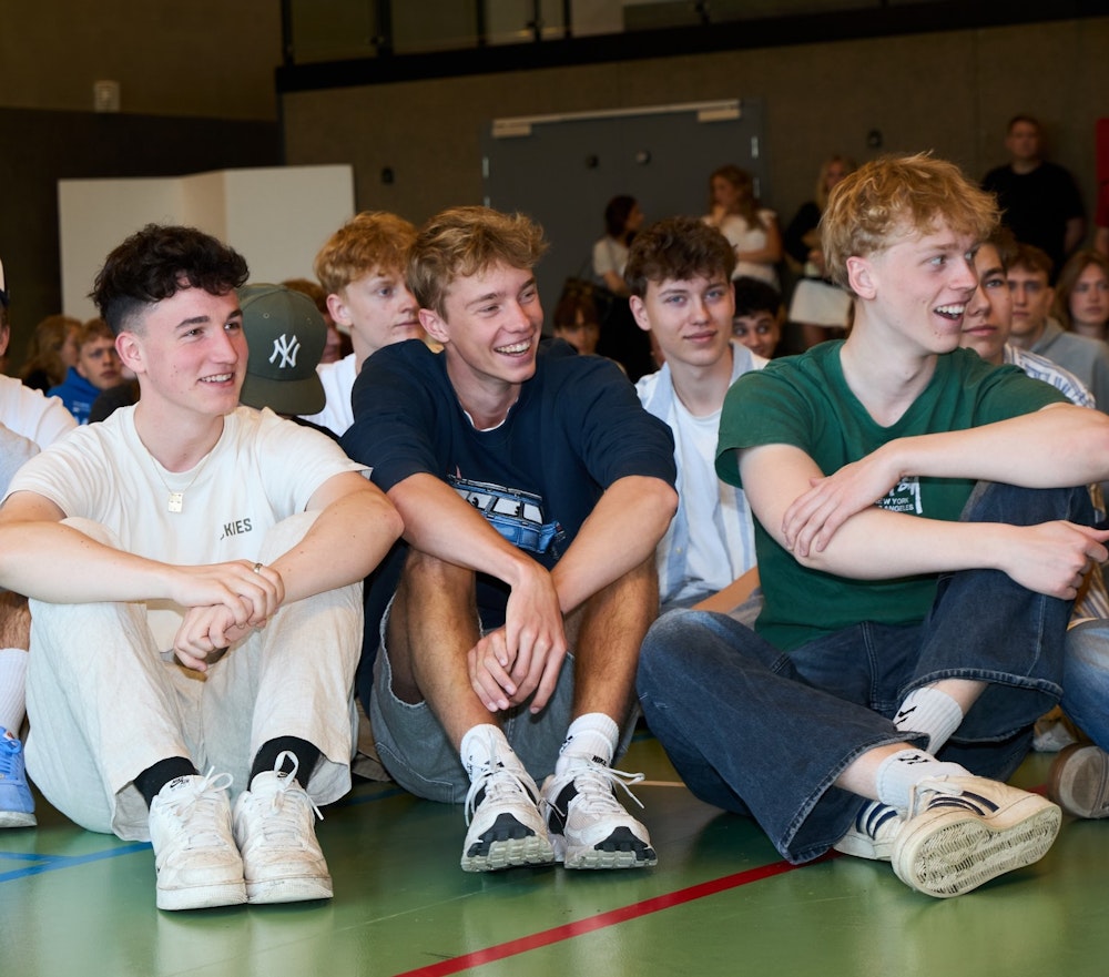 Grinende elever sidder på gulvet i skolens hal til morgensamling