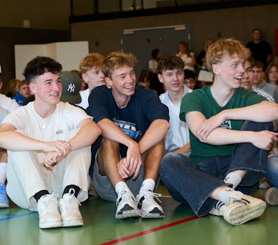 Grinende elever sidder på gulvet i skolens hal til morgensamling