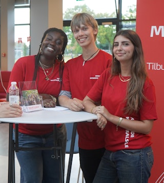 Halvtotal af tre elever i røde T-shirts, der står ved et bord til 1.g-informationsaften