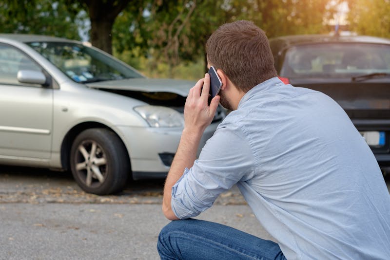Man sitting down talking on a cell phone with a car accident in the background.