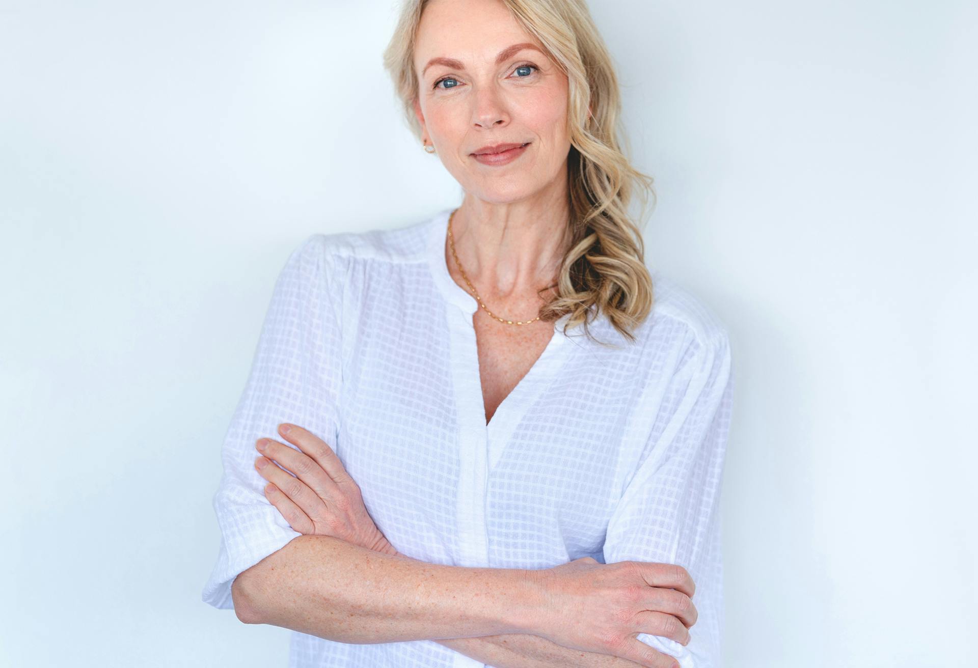 Woman in a white top with her arms crossed, against a wall