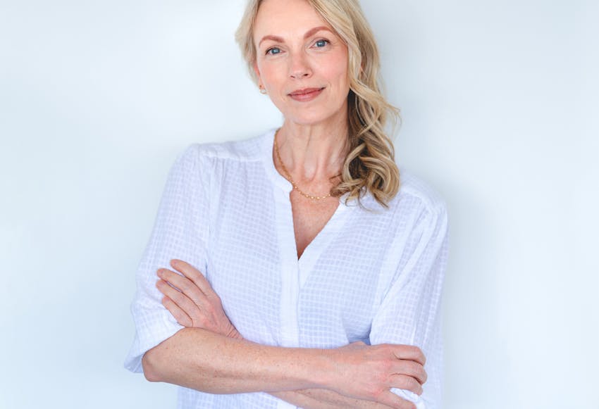 Woman in a white top with her arms crossed, against a wall