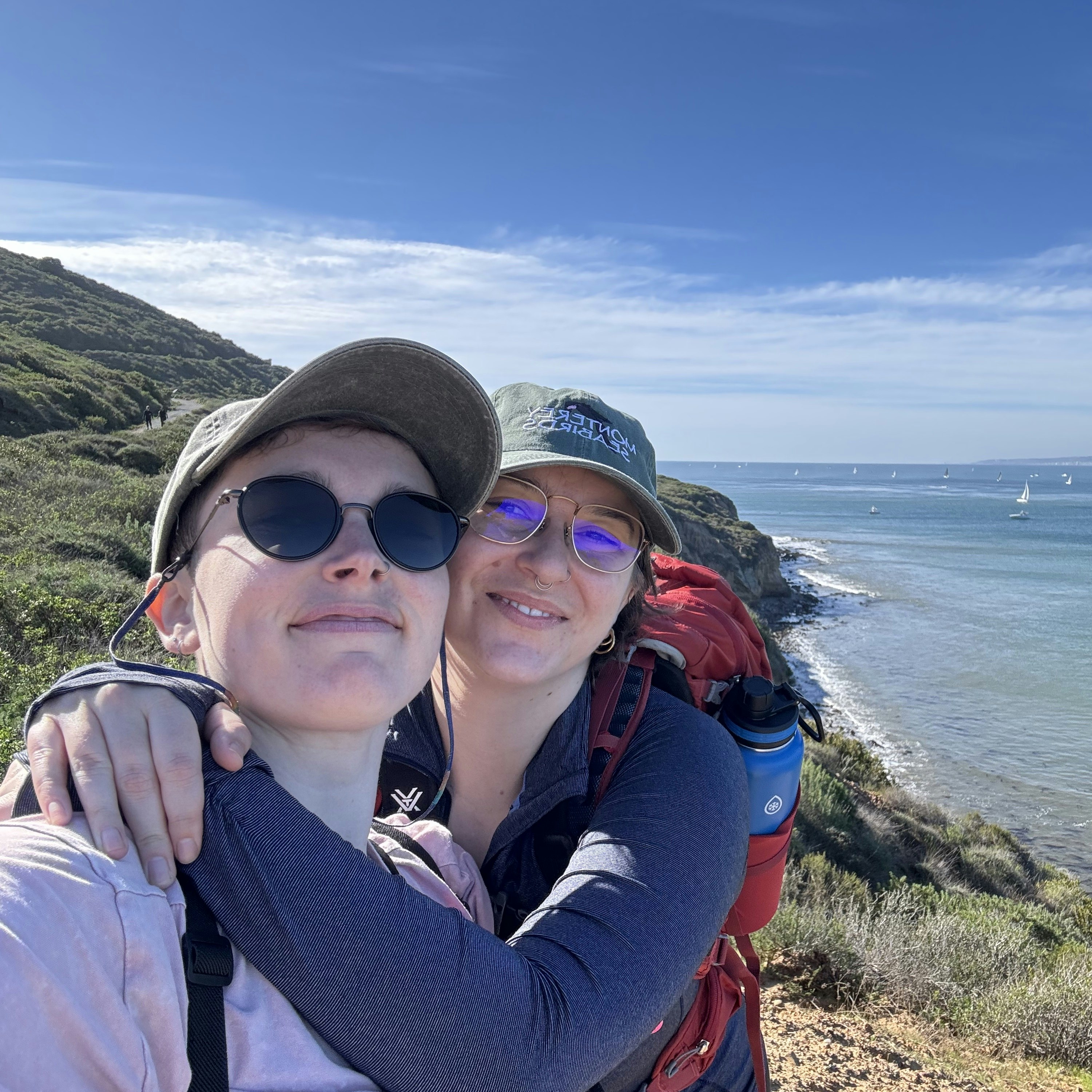 Erica, wearing a Monterey Seabirds hat, with her arms around her partner Tara and the ocean in the background.