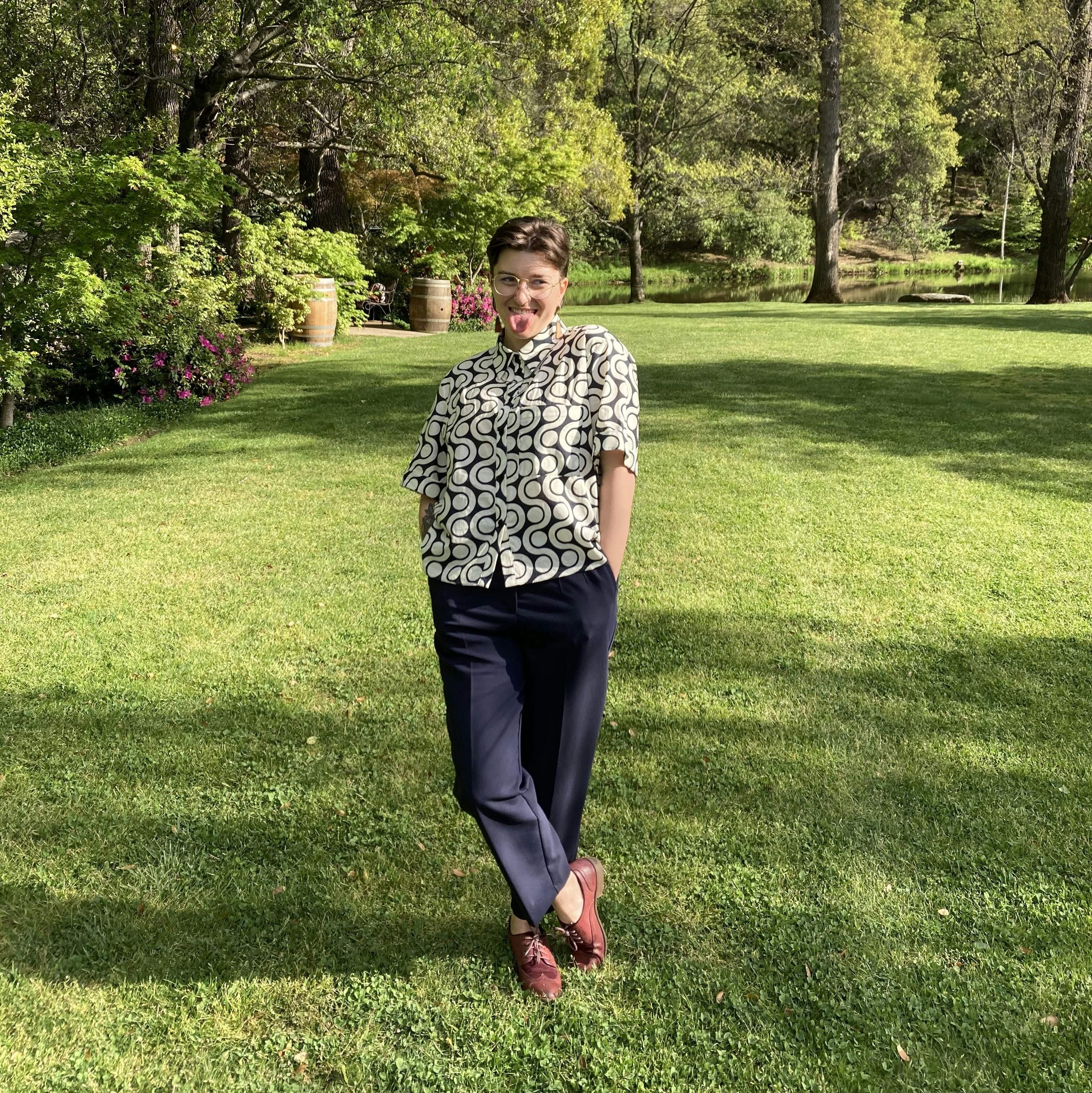 Erica wearing a white patterned shirt, blue slacks, and red oxfords with trees in the background. 