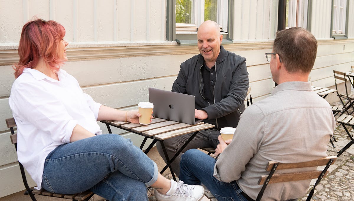 Tre av Midagons medarbetare sitter ner vid ett cafébord utomhus. En har sin dator framför sig och de andra två har varsin kaffemugg i handen.