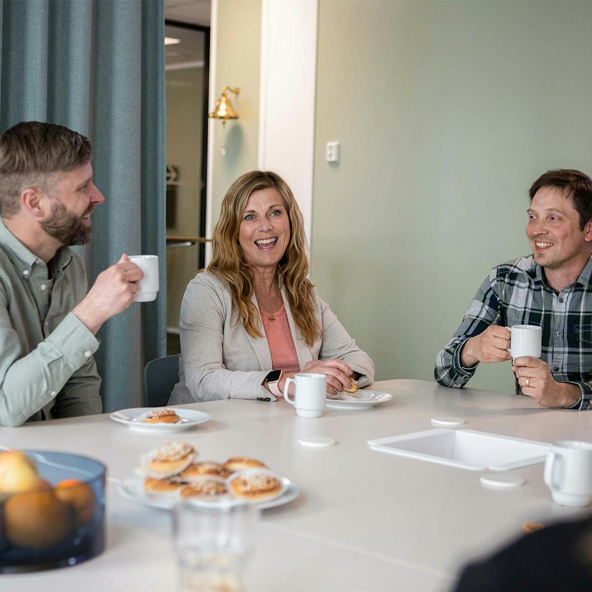 Tre kollegor fikar på Midagons Örebrokontor. De sitter ner vid ett bord med en varsin kaffekopp. På bordet står ett fat med bullar och två av kollegorna har en assiett framför sig med en varsin bulle på.