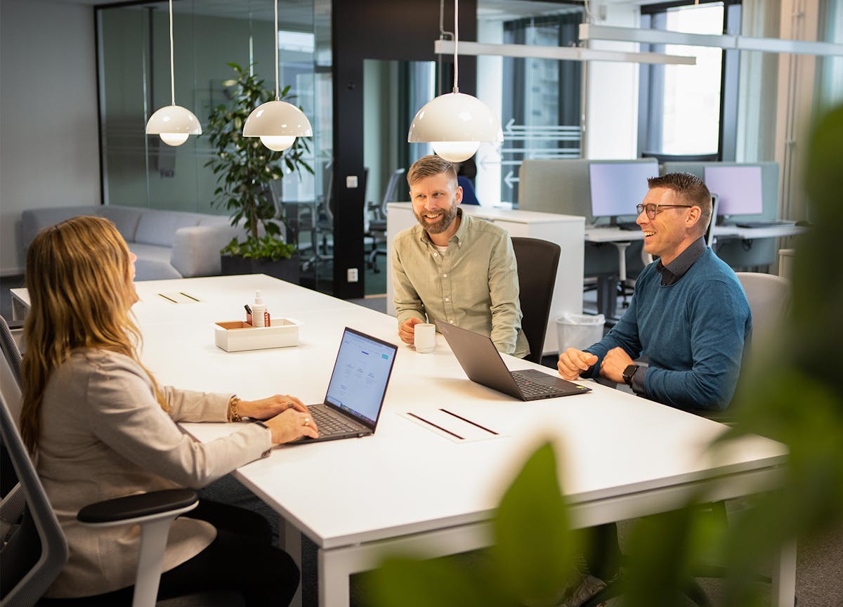 Tre kollegor sitter ner och arbetar vid ett bord på kontoret i Örebro. Två har sina datorer framför sig och den tredje sitter med en kaffekopp i handen. De tittar på varandra och ler.