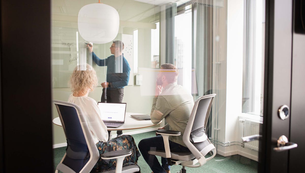 Tre kollegor arbetar i ett av konferensrummen på Midagons Örebrokontor. En av dem skriver på whiteboardtavlan på väggen och de andra två sitter ner vid bordet med sina datorer.