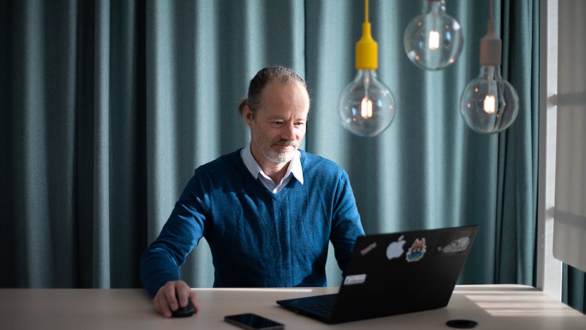 Bild på man som sitter vid en svart laptop och arbetar. Mannen har en blå tröja och vit skjorta under.