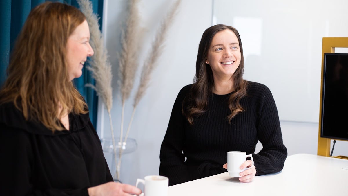 Emelie Fredriksson, konsult på Midagon Sverige. Brunhårig, svartklädd kvinna dricker kaffe med en kollega på Midagon Sveriges kontor. Båda ler.